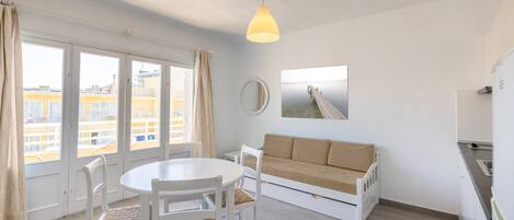 A bright room with a dining table, white chairs, a sofa, a hanging pendant light, a round mirror, and a view of buildings through large windows.