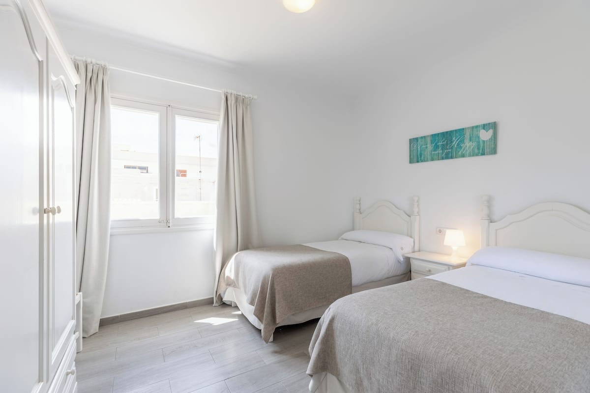 A hotel room with two beds, a window with curtains, a bedside table with a lamp, and a wall-mounted artwork.