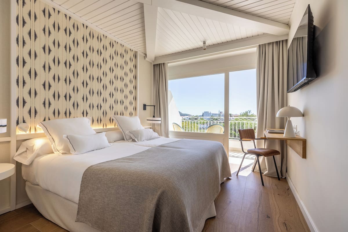 A modern hotel room with a large bed, a desk, a chair, a TV, and a balcony with a city view.