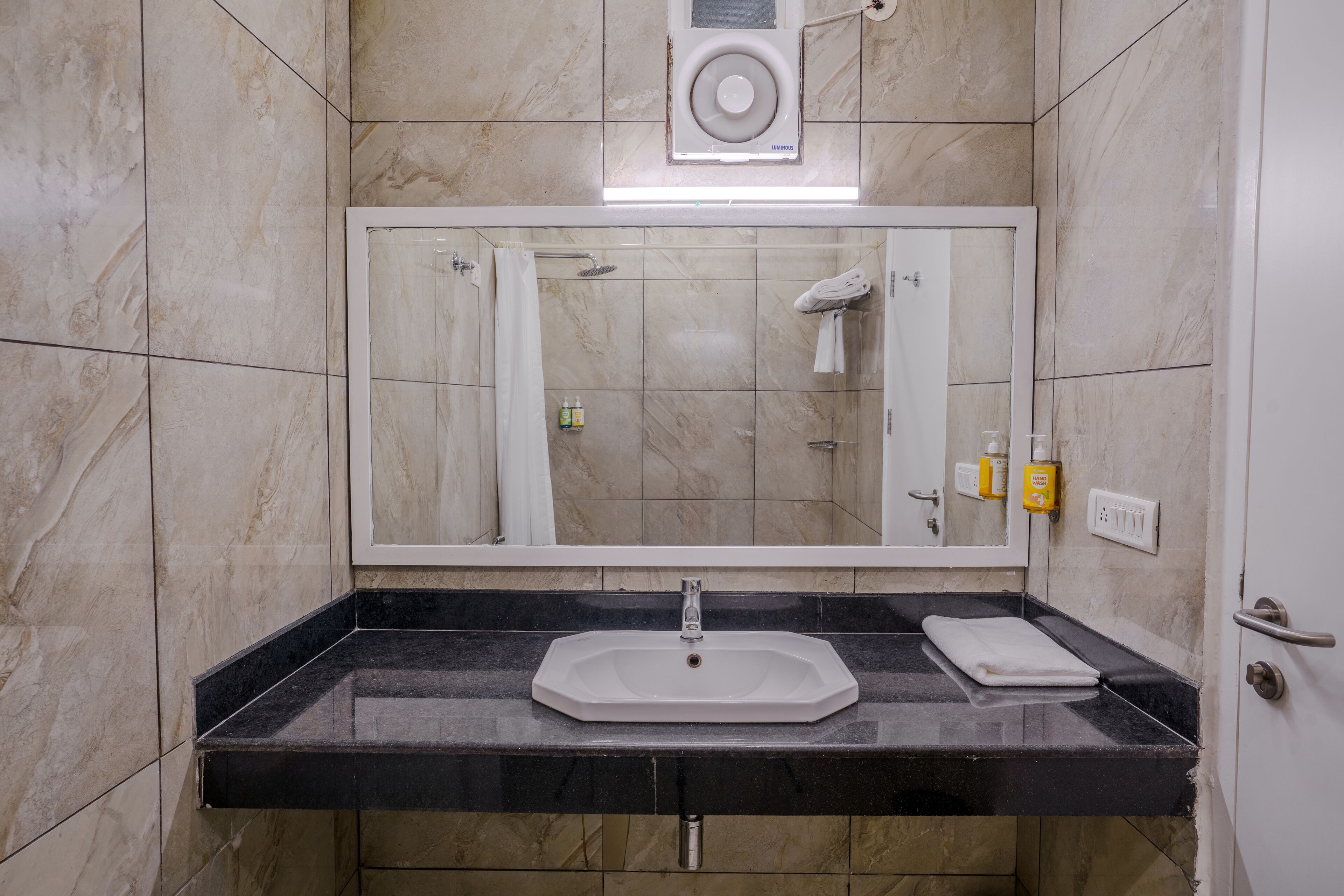 Une salle de bain avec un mur recouvert de carreaux de marbre, un plan de toilette noir, un lavabo blanc, un miroir, une serviette et une douche.