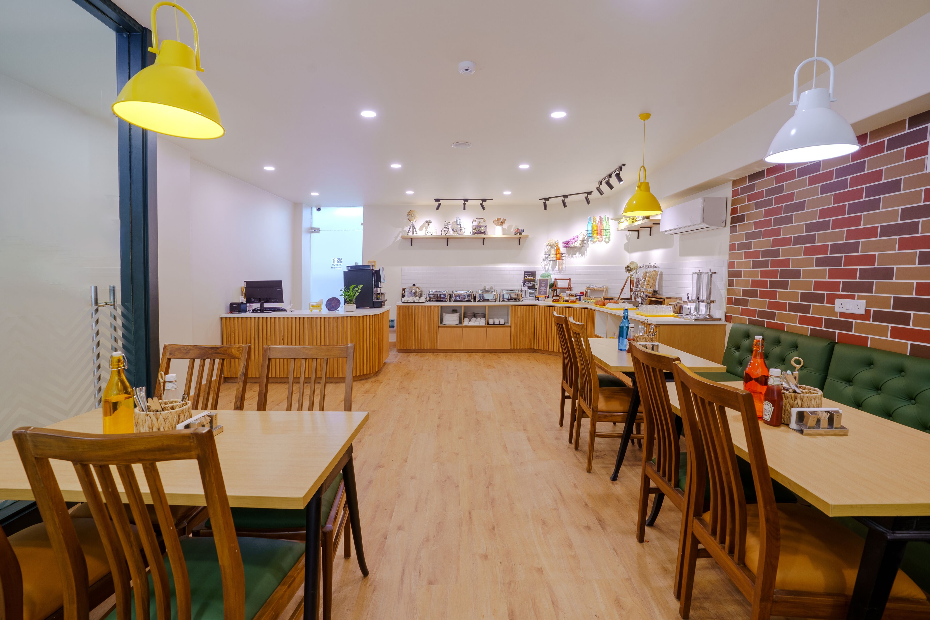 Un comedor moderno con mesas y sillas de madera, un rincón con asientos tipo banca en tono verde, y una cocina al fondo.