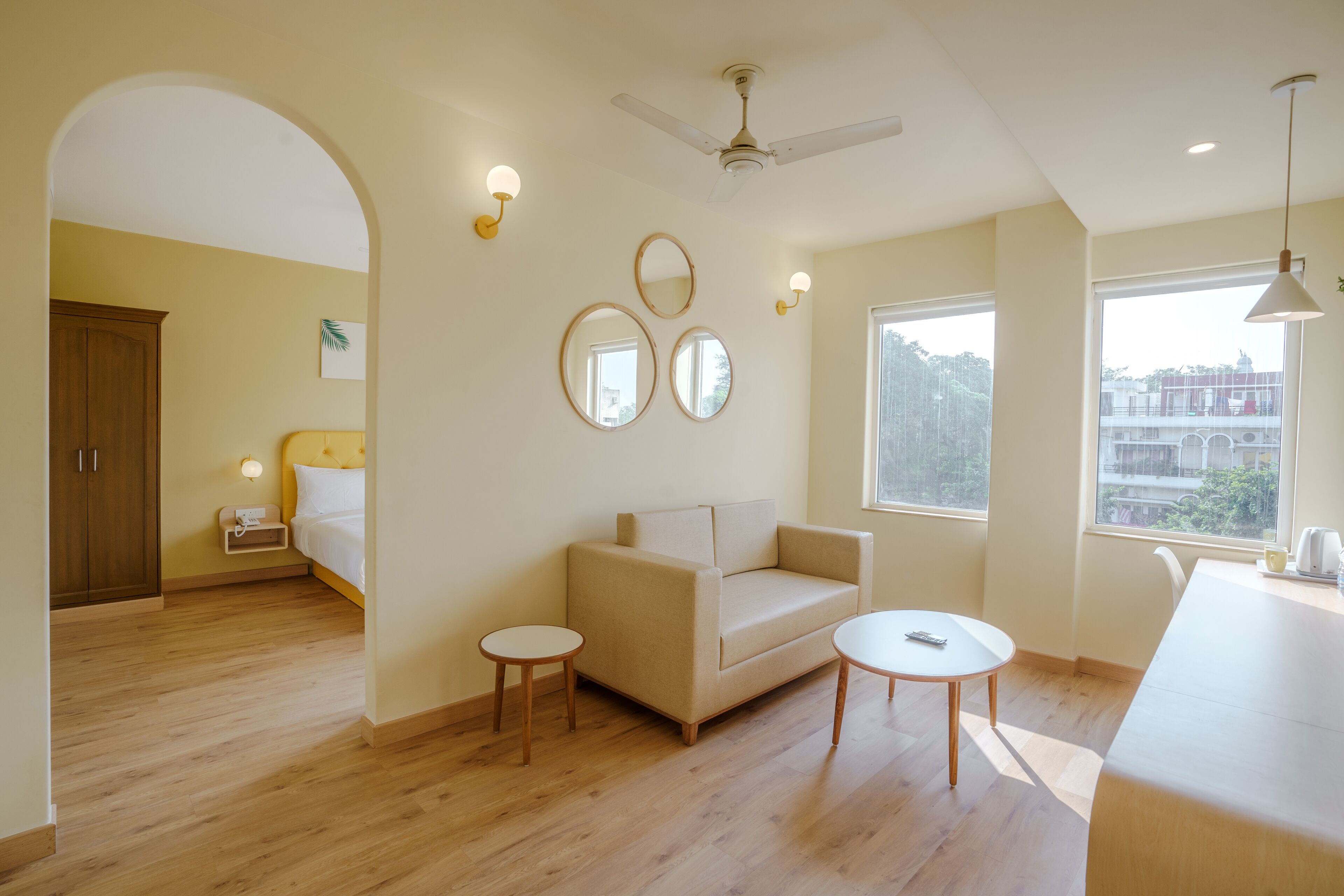 Een moderne woonkamer met een bank, twee bijzettafeltjes, een plafondventilator en grote ramen met uitzicht op gebouwen en bomen.