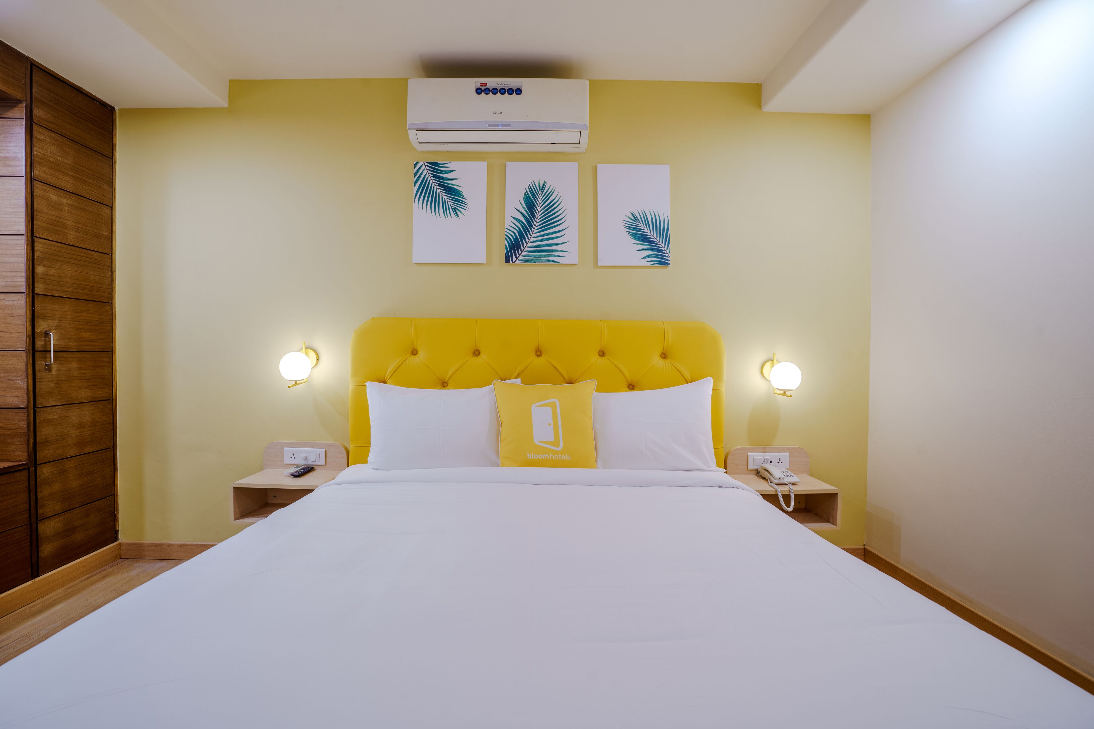 A hotel room with a yellow headboard, white bedding, a bedside telephone, and wall-mounted air conditioning.