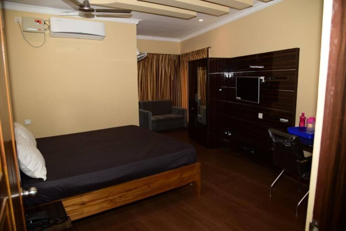 A bedroom with a bed, a chair, a desk, a TV, and an air conditioning unit.