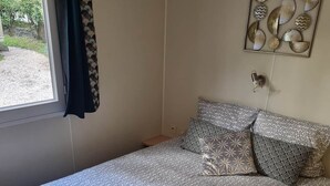 A bedroom with a bed, patterned bedding, a window with curtains, and a wall decoration. - Mobile home Trigano 5 people AIR-CONDITIONED (ST JEAN DE VALERISCLE)