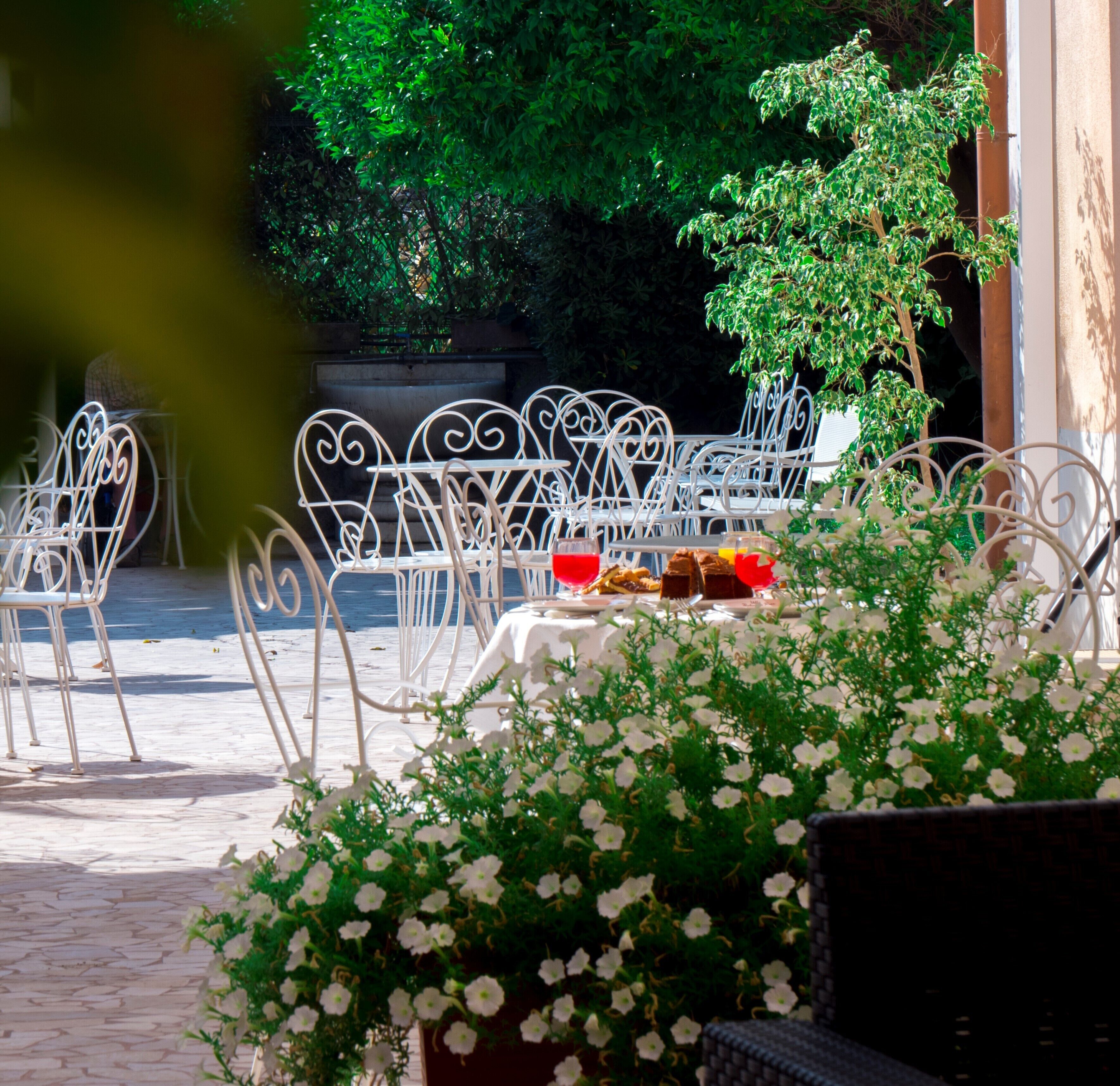Een terras met witte metalen stoelen en tafels, omgeven door groen en bloemen.