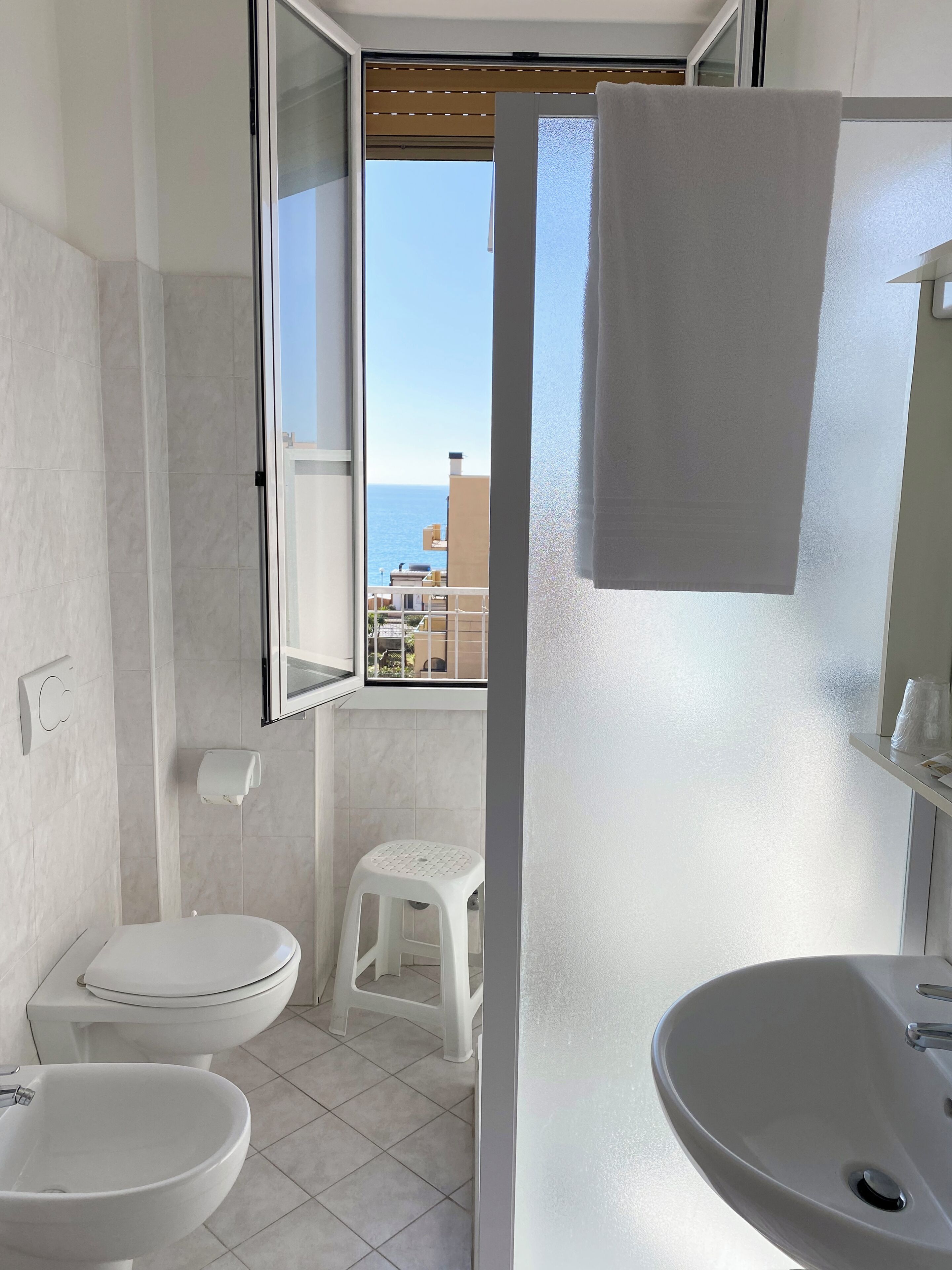 A bathroom with a toilet, bidet, and sink, a small stool, a towel hanging on the wall, and a window with a view of buildings and the sea.
