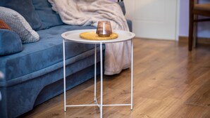 En blå sofa med pude, et lille hvidt bord med en genstand af glas, og trægulv.