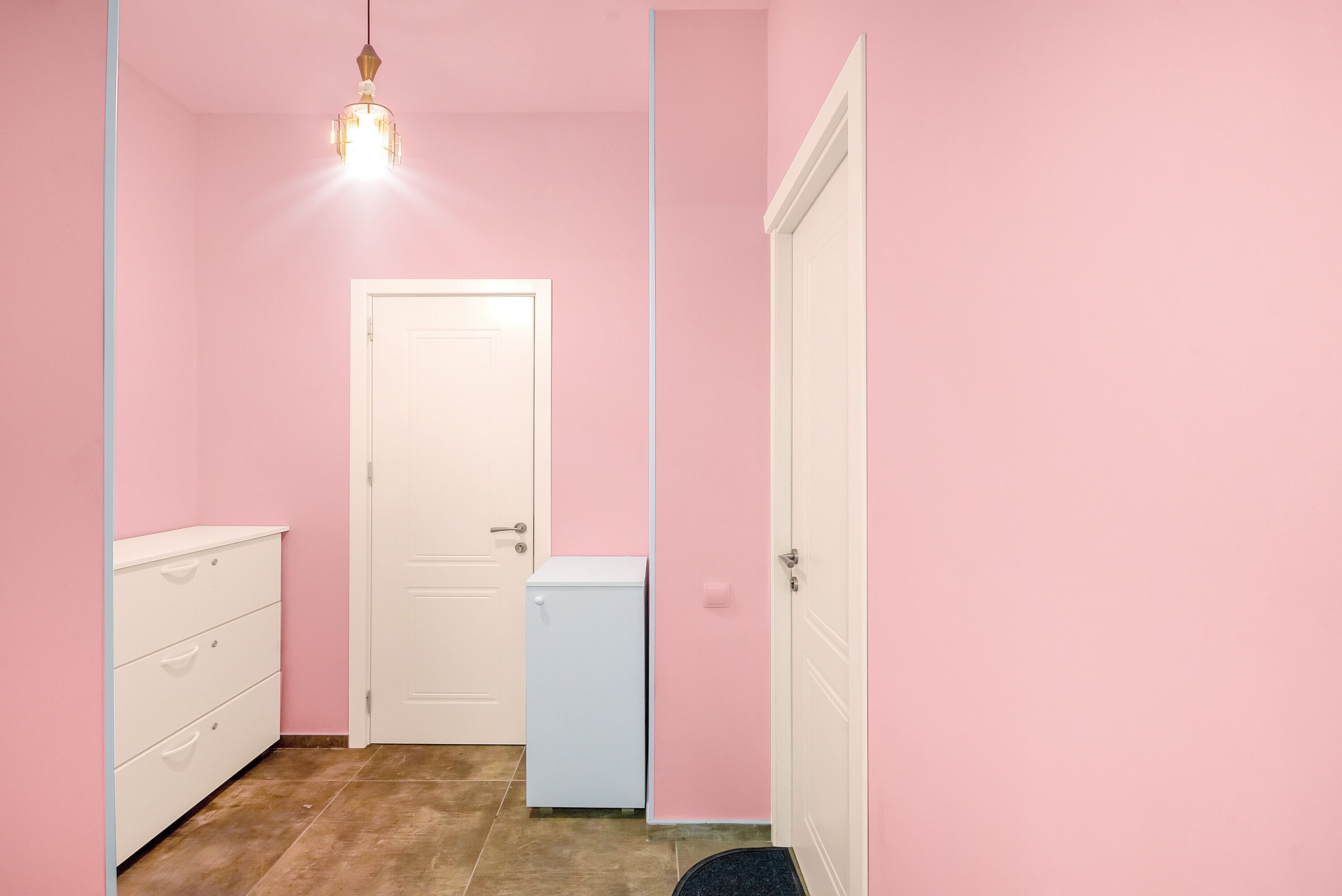 A small, pink-walled room with a white door, a white dresser, and a small white appliance.