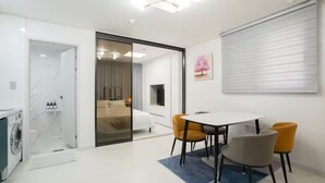 A modern hotel room with a glass door leading to a bathroom, a dining area with a table and chairs, and a large window with blinds.