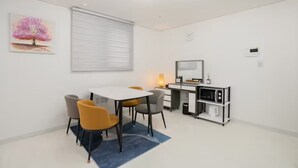 A modern, minimalist room with a white color scheme, featuring a dining table with four chairs, a small desk with a computer, and a large window with blinds.