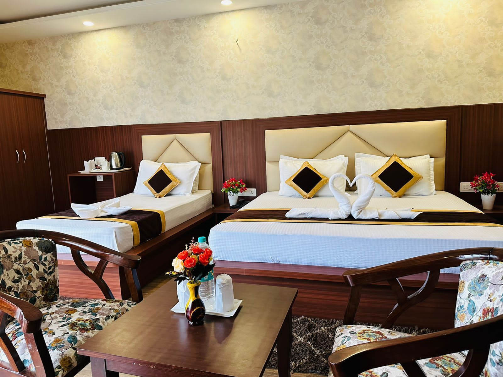Ein Hotelzimmer mit zwei Betten, einem Holztisch, floralen Stühlen und einer Vase mit Blumen.