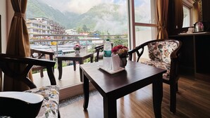 Une pièce dotée d’une grande fenêtre donnant sur un paysage montagneux et sur une ville située en contrebas. On y trouve une table en bois sur laquelle sont posés un vase de fleurs et une bouteille d’eau.