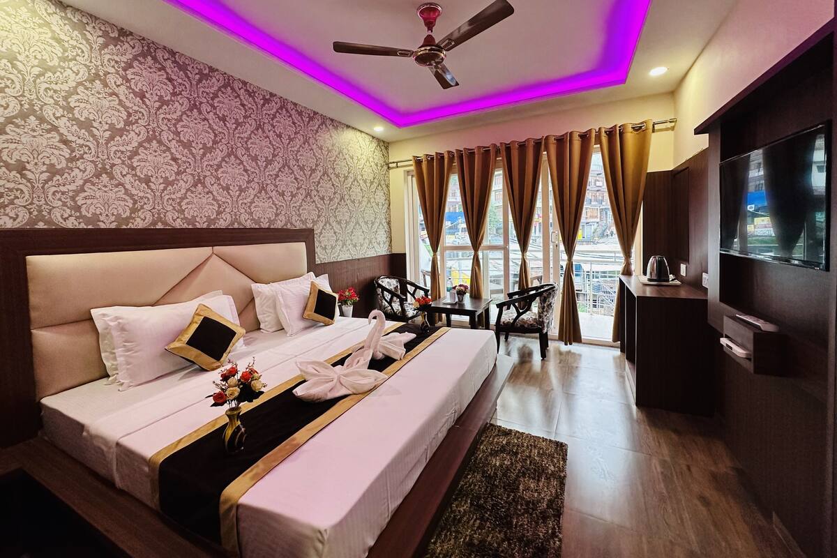A hotel room with a large bed, a ceiling fan, a TV, and a view of the city through the windows.