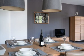 Comedor con mesa de madera dispuesta para cuatro, una botella de vino y un televisor al fondo.