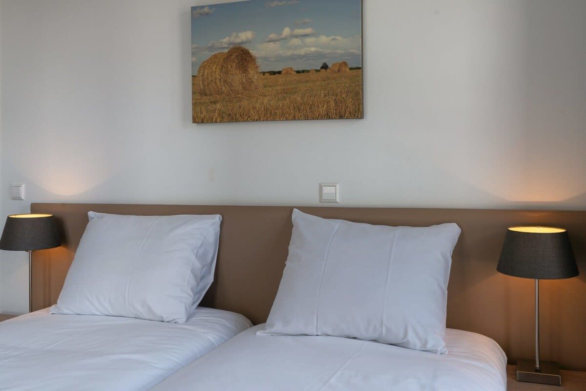 Une chambre d’hôtel avec deux lits, une tête de lit et un tableau accroché au mur représentant un champ avec des bottes de foin.