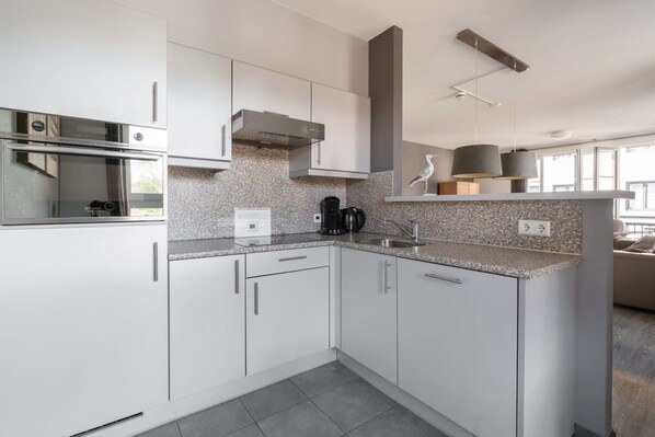 A modern kitchen with white cabinetry, a built-in oven, a coffee maker, and a pendant light. - Apartment in Cadzand-Bad | Duinhof V 1-7 (Cadzand)