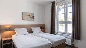 A double bed with white linens, a nightstand with a lamp, a window with curtains, and a wall-mounted artwork. - Apartment in Cadzand-Bad | Duinhof V 1-7 (Cadzand)