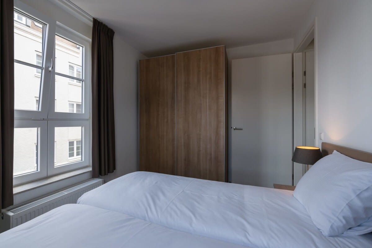 Ein Hotelzimmer mit Bett, Kleiderschrank, Fenster mit Vorhängen und einer Nachttischlampe.