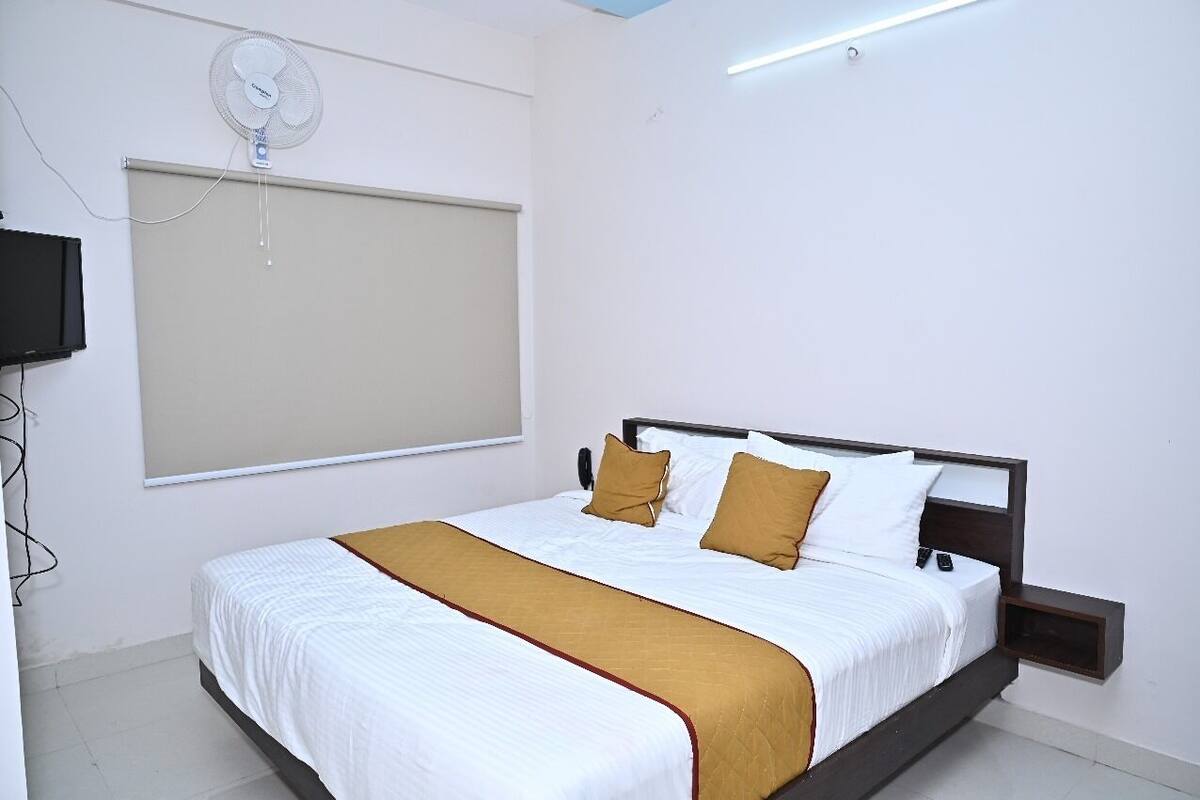A neatly made bed with white and brown bedding, a wall-mounted fan, and a TV on the left.
