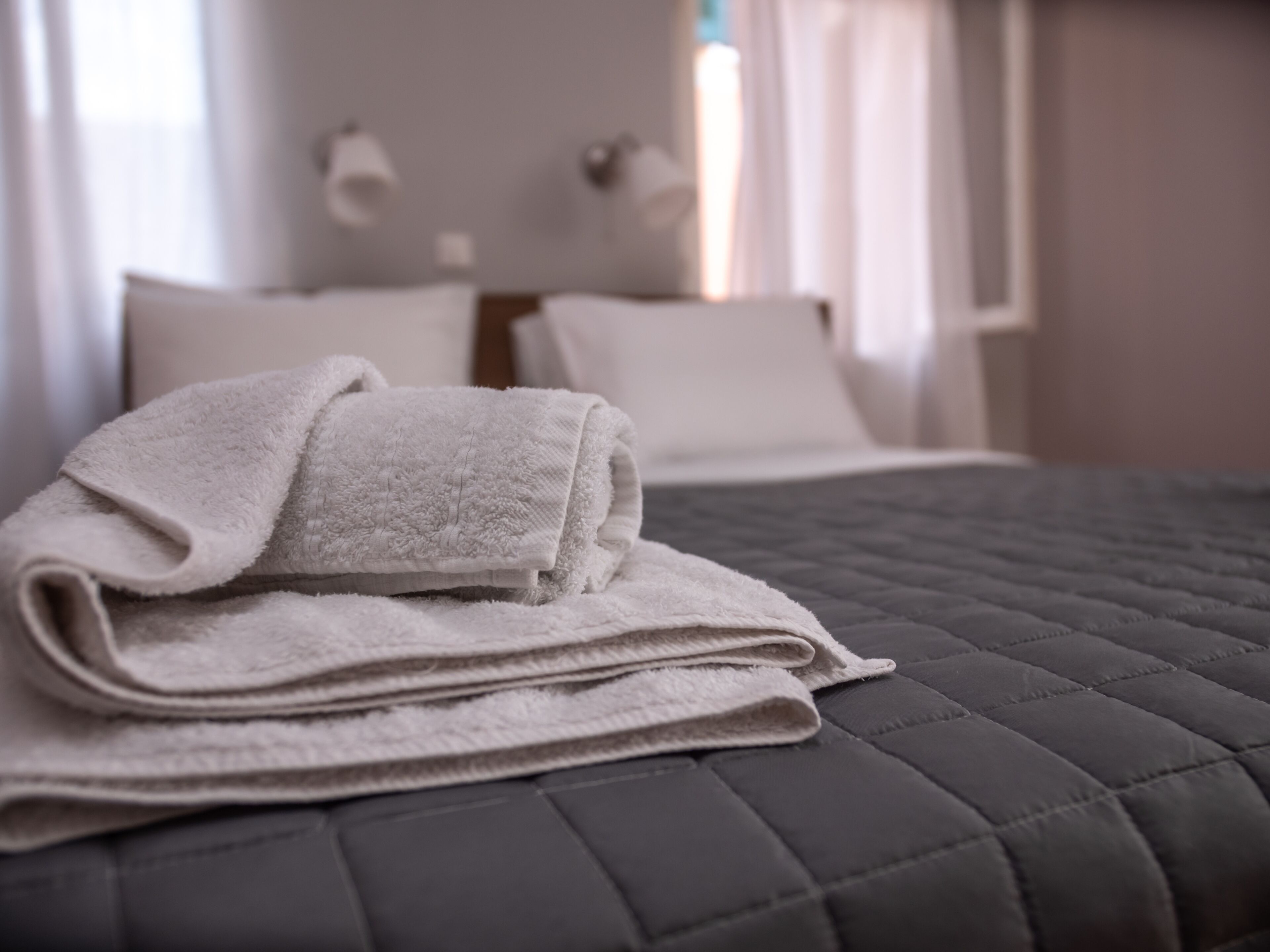 Un lit bien fait, avec une serviette blanche pliée posée sur le dessus, une couvre-lit matelassée de couleur sombre et une tête de lit agrémentée de lampes fixées au mur.