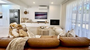 A modern living room with a sofa, a coffee table, and a TV. There is a large window with blinds, a kitchen area with a refrigerator, and a fireplace.