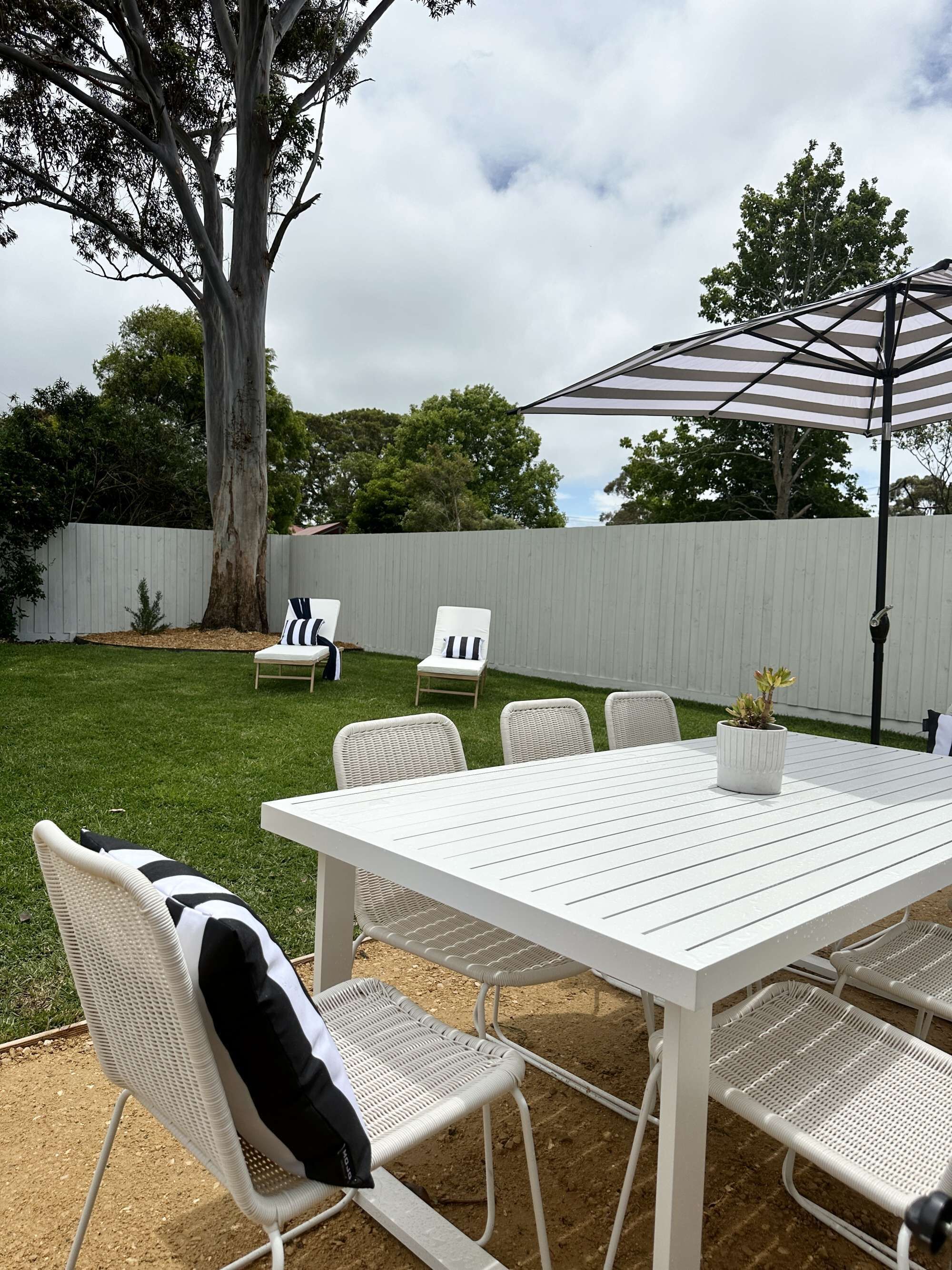 Un patio con una mesa y sillas blancas, un parasol a rayas y un árbol.