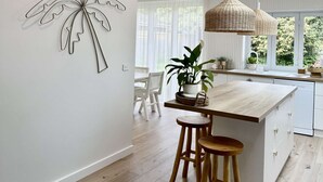 A modern kitchen with a central island, wooden stools, pendant lighting, and a large window with a view of greenery.