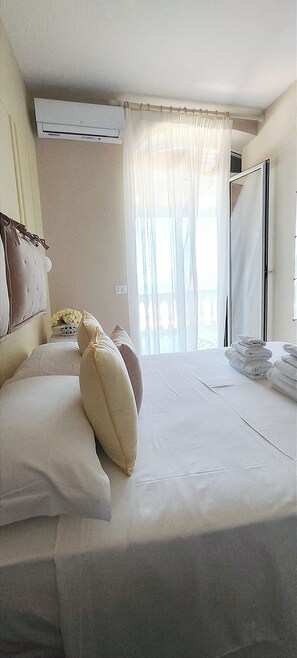 A hotel room with a bed, a window with sheer curtains, an air conditioning unit, and a stack of towels. - Villa Antonietta Ischia  (Forio)