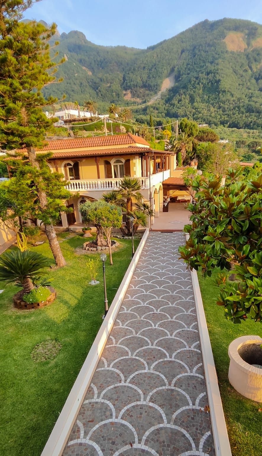 Uma trilha conduz a um edifício com telhado de azulejos, cercado por vegetação e montanhas ao fundo.