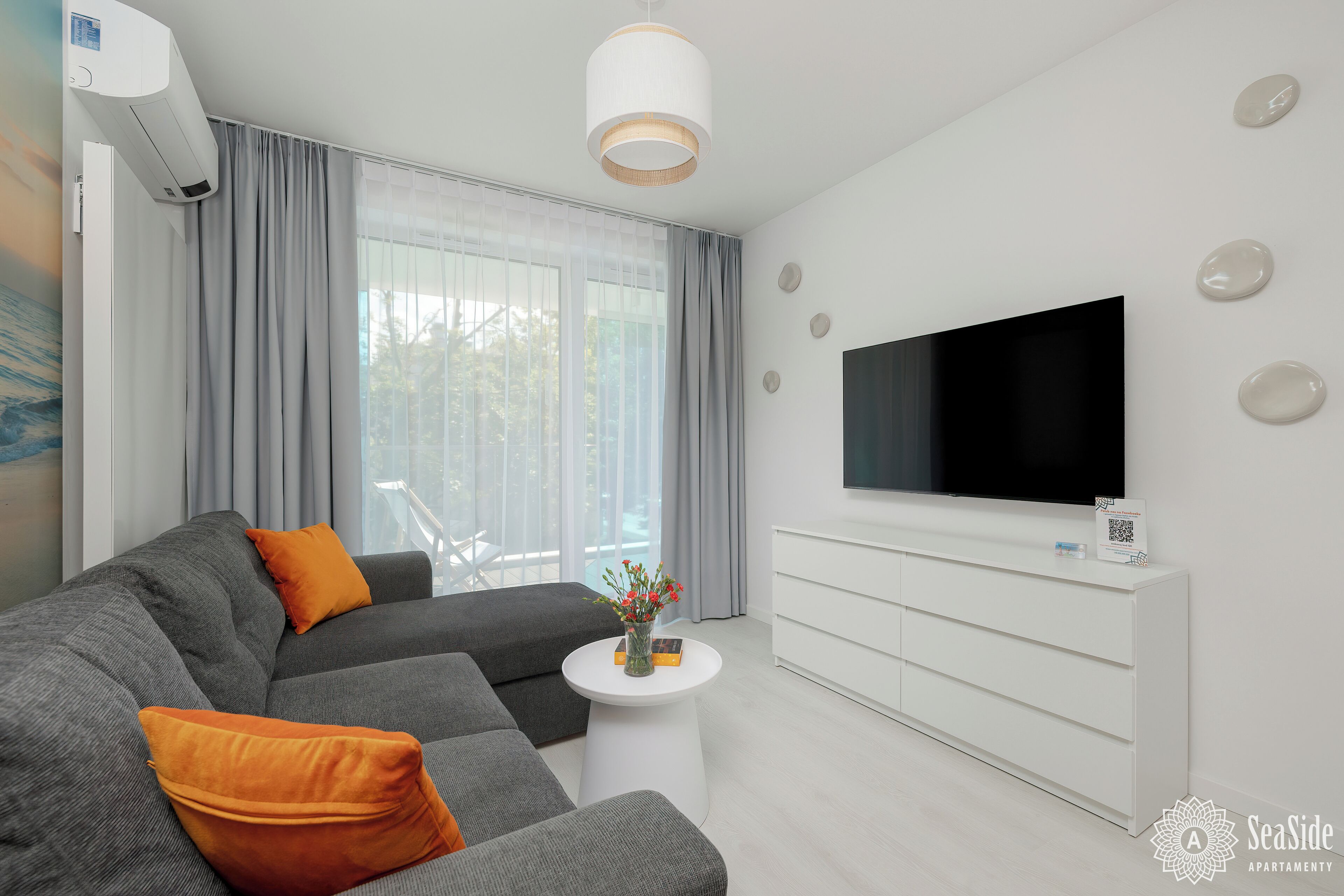 A modern living room with a grey sofa, orange cushions, a white coffee table with a vase of flowers, a flat-screen TV on a white cabinet, and a large window with grey curtains.