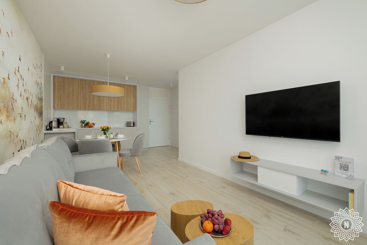 Ett modernt vardagsrum med en grå soffa, ett träbord med frukt på, en platt-TV på väggen och ett kök i bakgrunden.