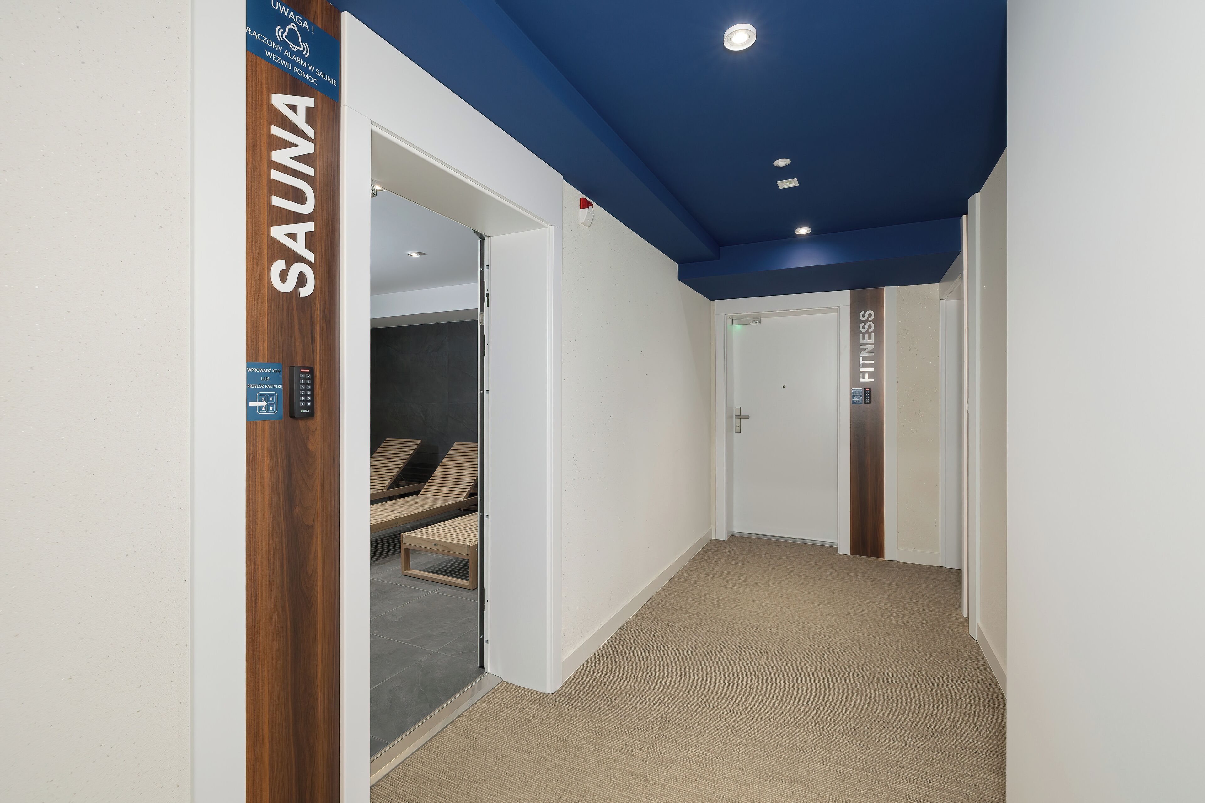 A hallway leading to a sauna area with a wooden sign, a control panel, and a door labeled "FITNESS".