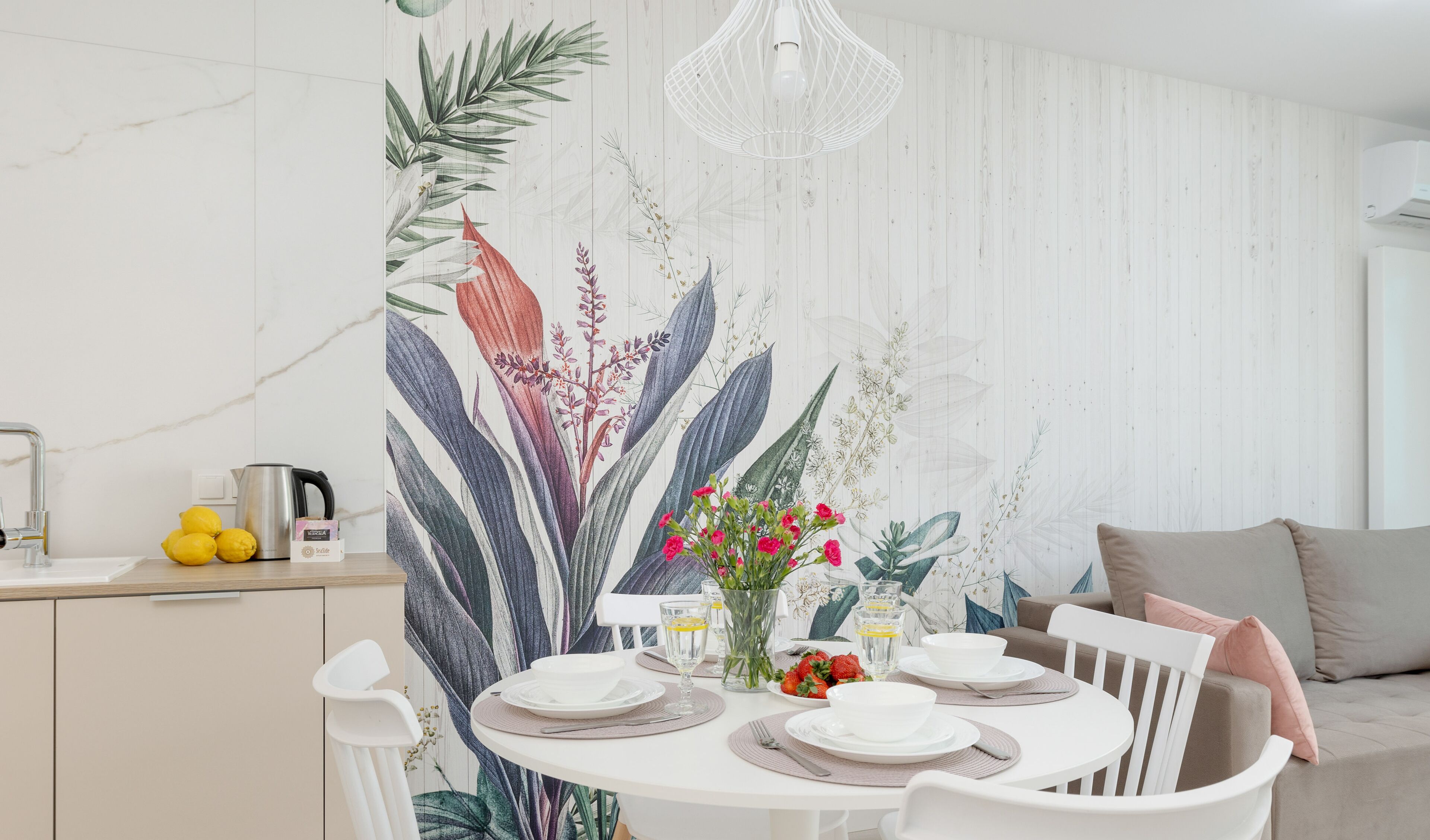 Un coin repas avec une table ronde dressée pour quatre personnes, des chaises blanches, un luminaire suspendu blanc et un mur orné d’un papier peint à motifs végétaux.