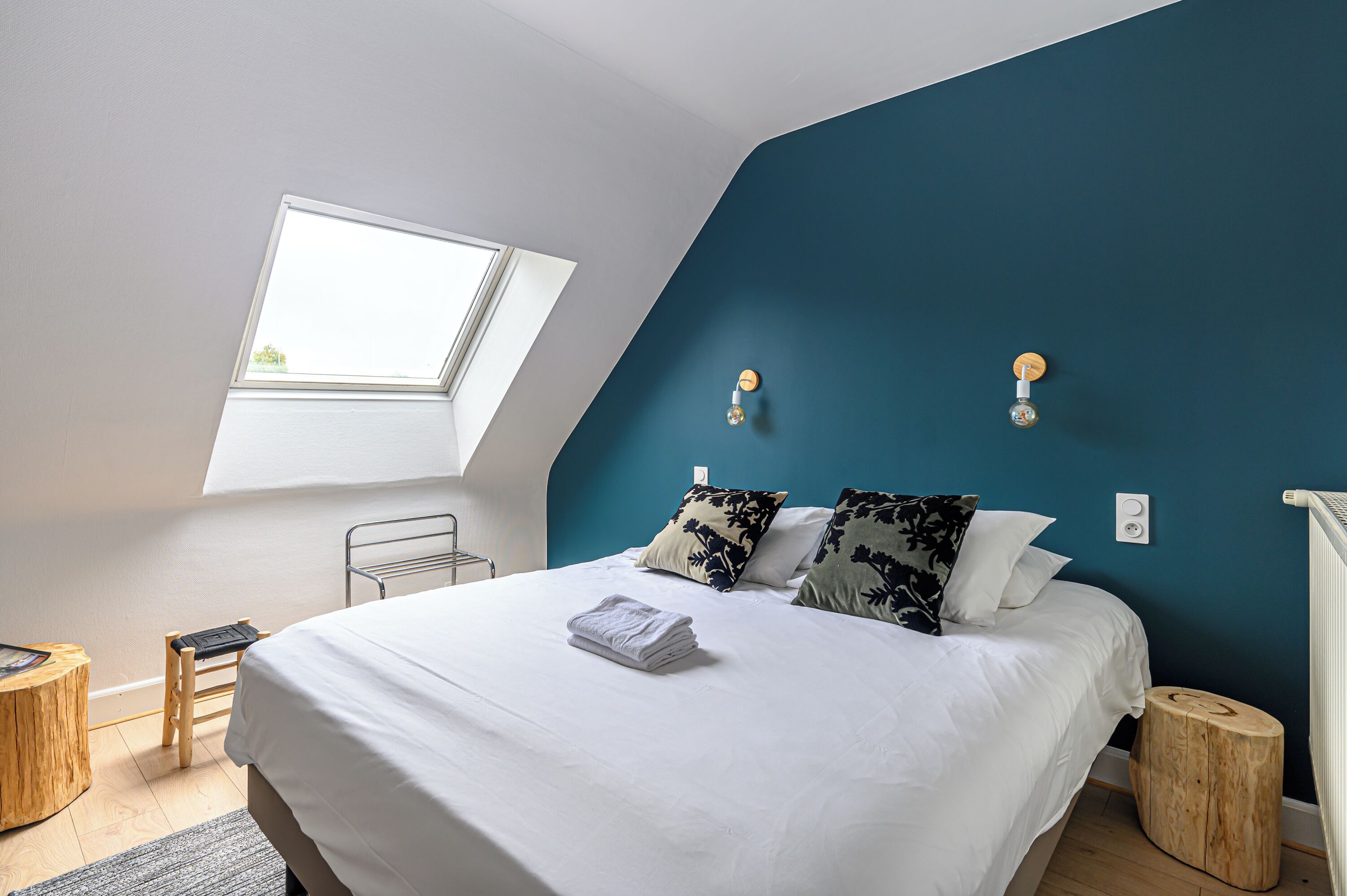 A bedroom with a skylight, a bed with white linens, two pillows with floral patterns, and a wooden stool.