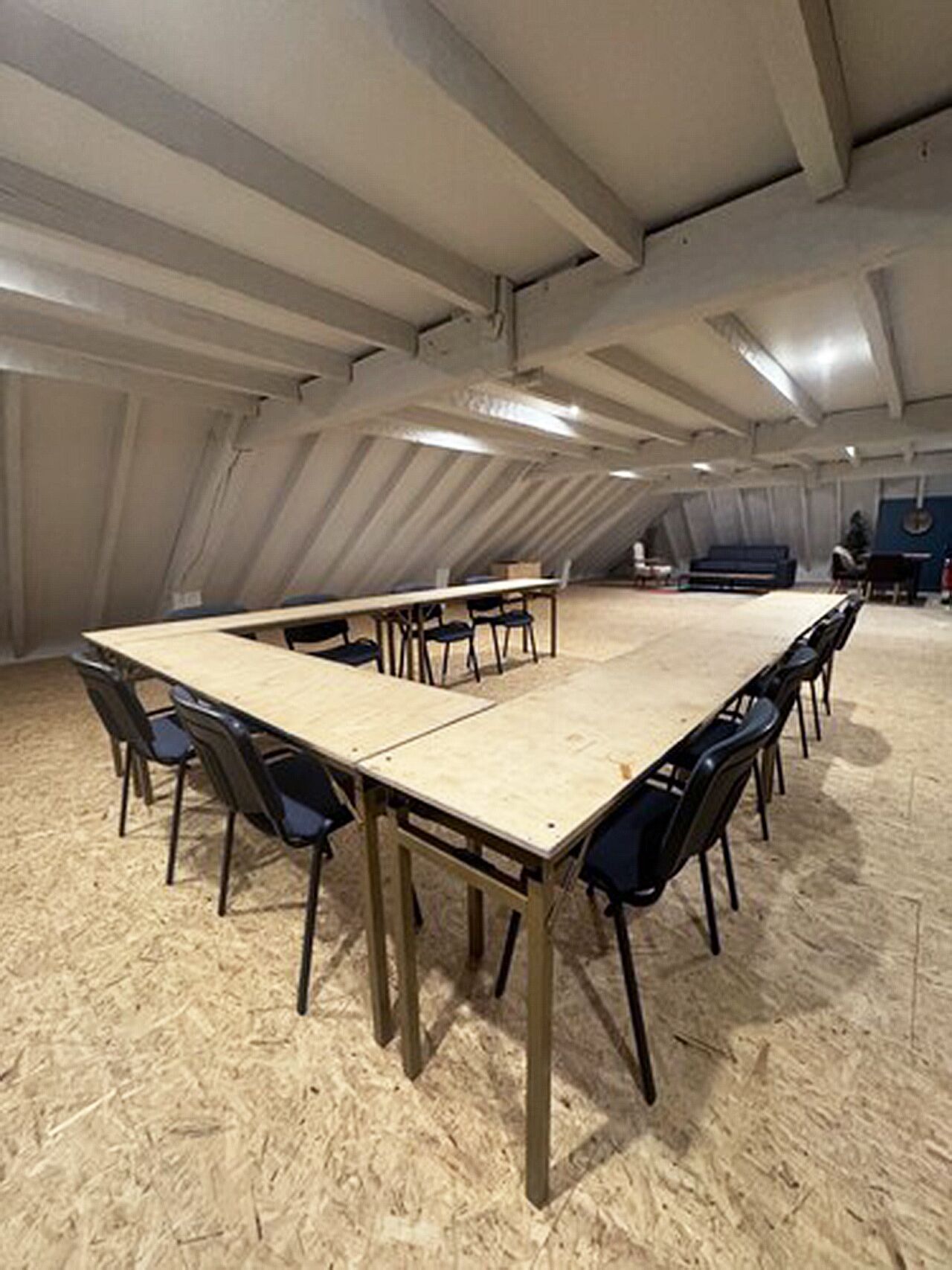 A large, open room with a high ceiling and exposed beams, featuring a long wooden table surrounded by chairs, a blue sofa, and a small table with chairs.