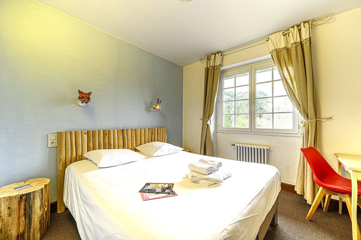 Une chambre d’hôtel avec un lit, une chaise rouge, une table de chevet en bois et une fenêtre avec des rideaux.