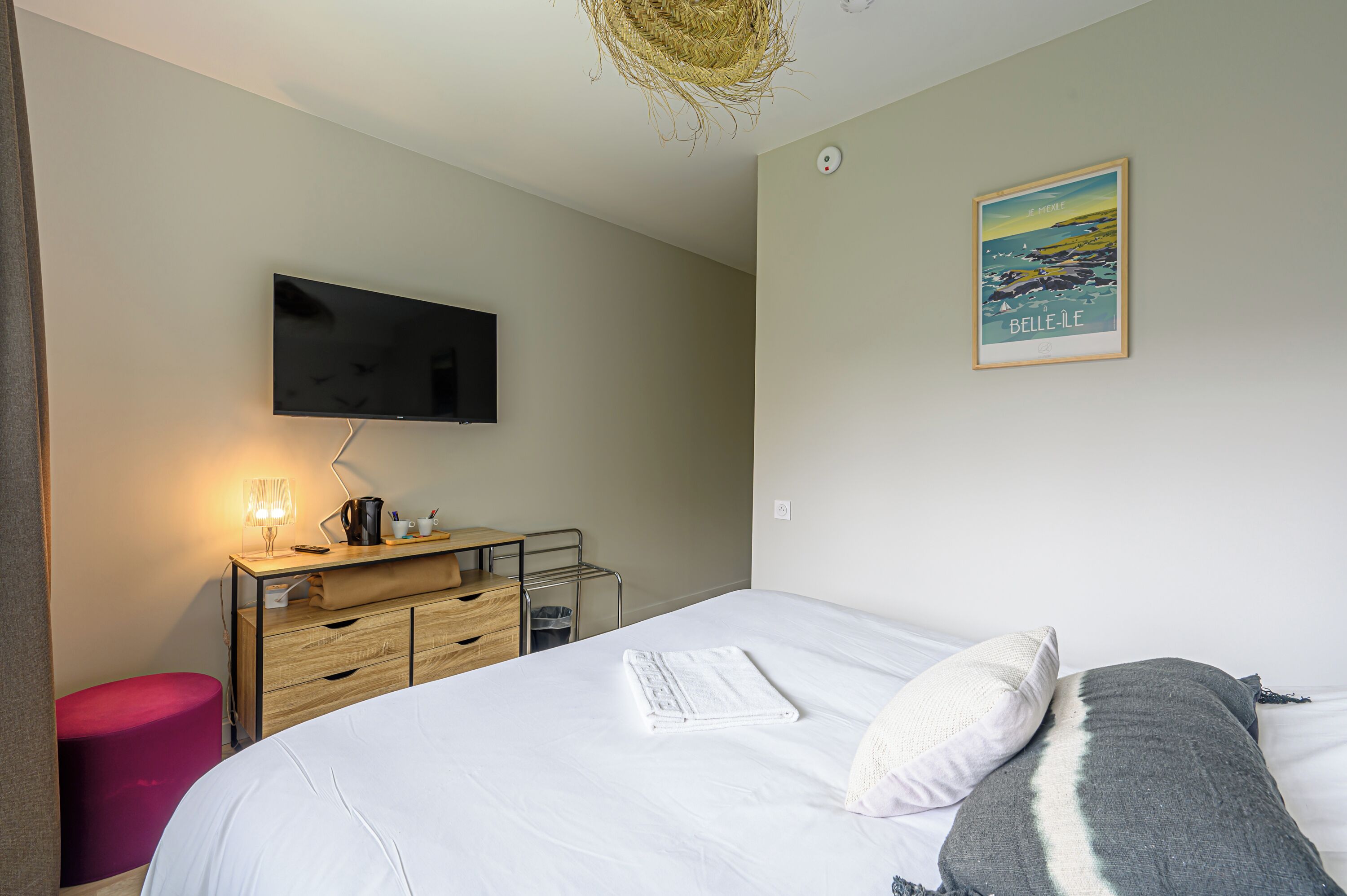 A hotel room with a bed, a desk with a lamp, a flat-screen TV, a wooden dresser, and a framed picture on the wall.