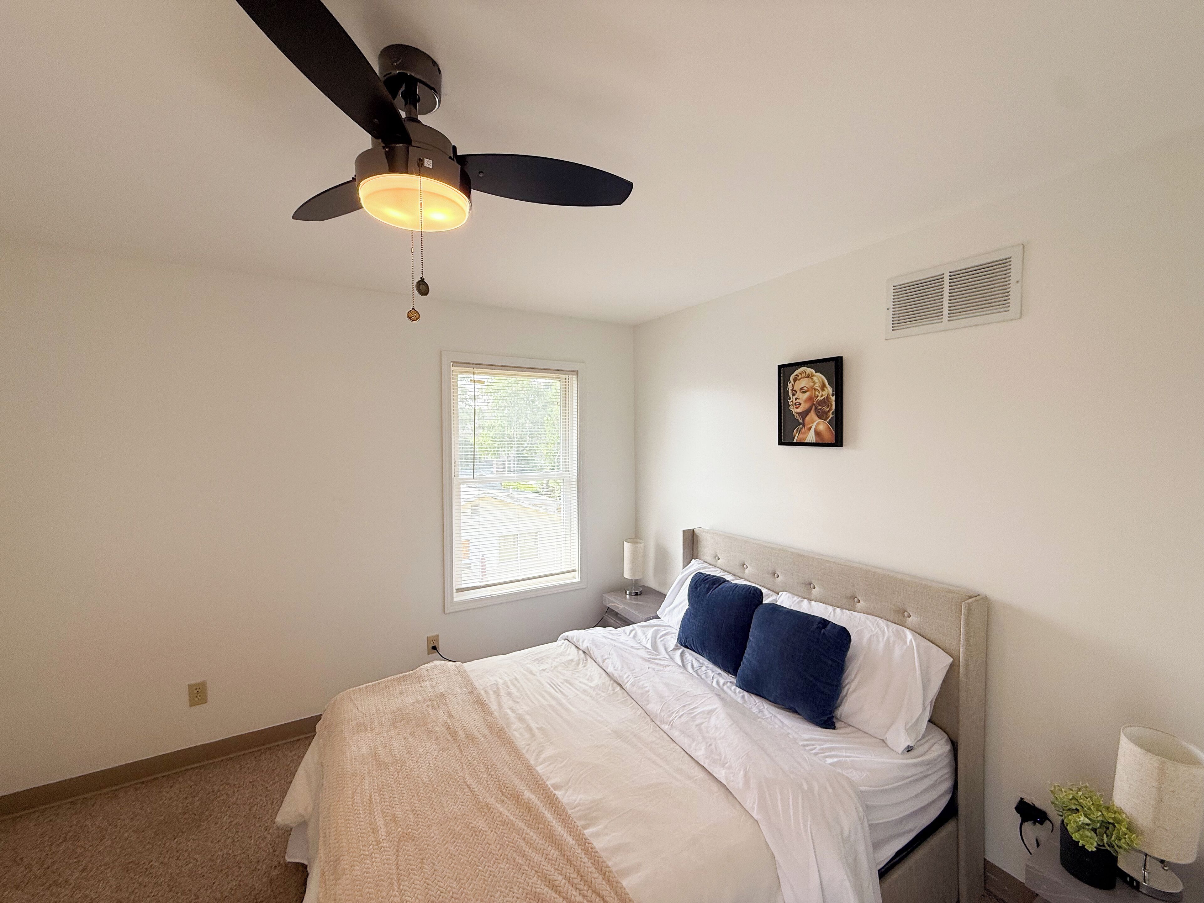 Quarto com cama, ventilador de teto, janela, ventilação na parede e um quadro emoldurado.