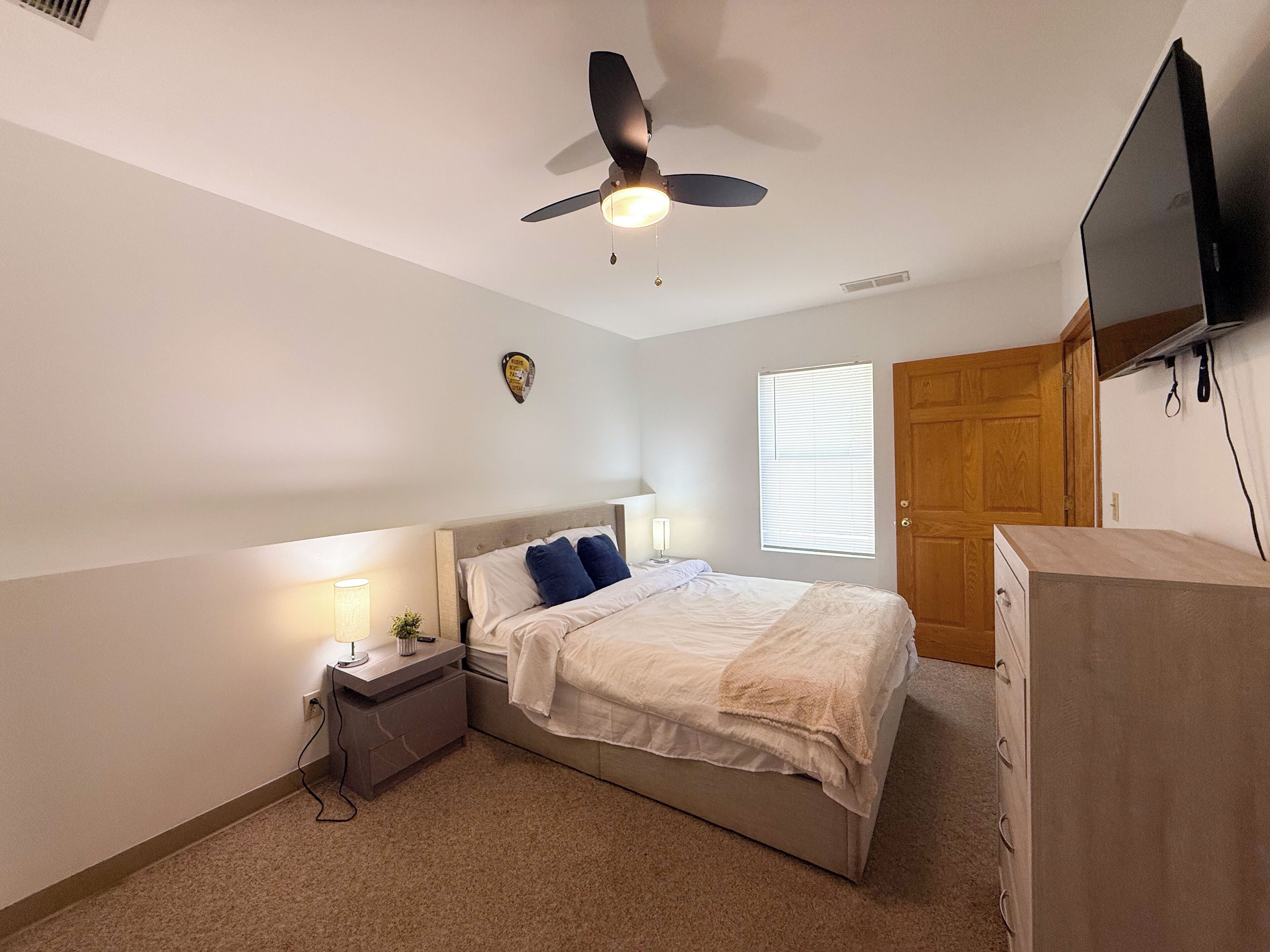 A bedroom with a bed, nightstand, lamp, ceiling fan, window, and a TV mounted on the wall.