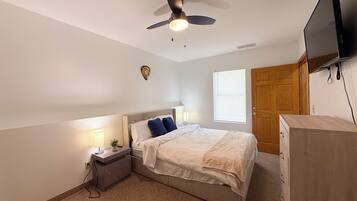 A bedroom with a bed, nightstand, lamp, ceiling fan, window, and a TV mounted on the wall.
