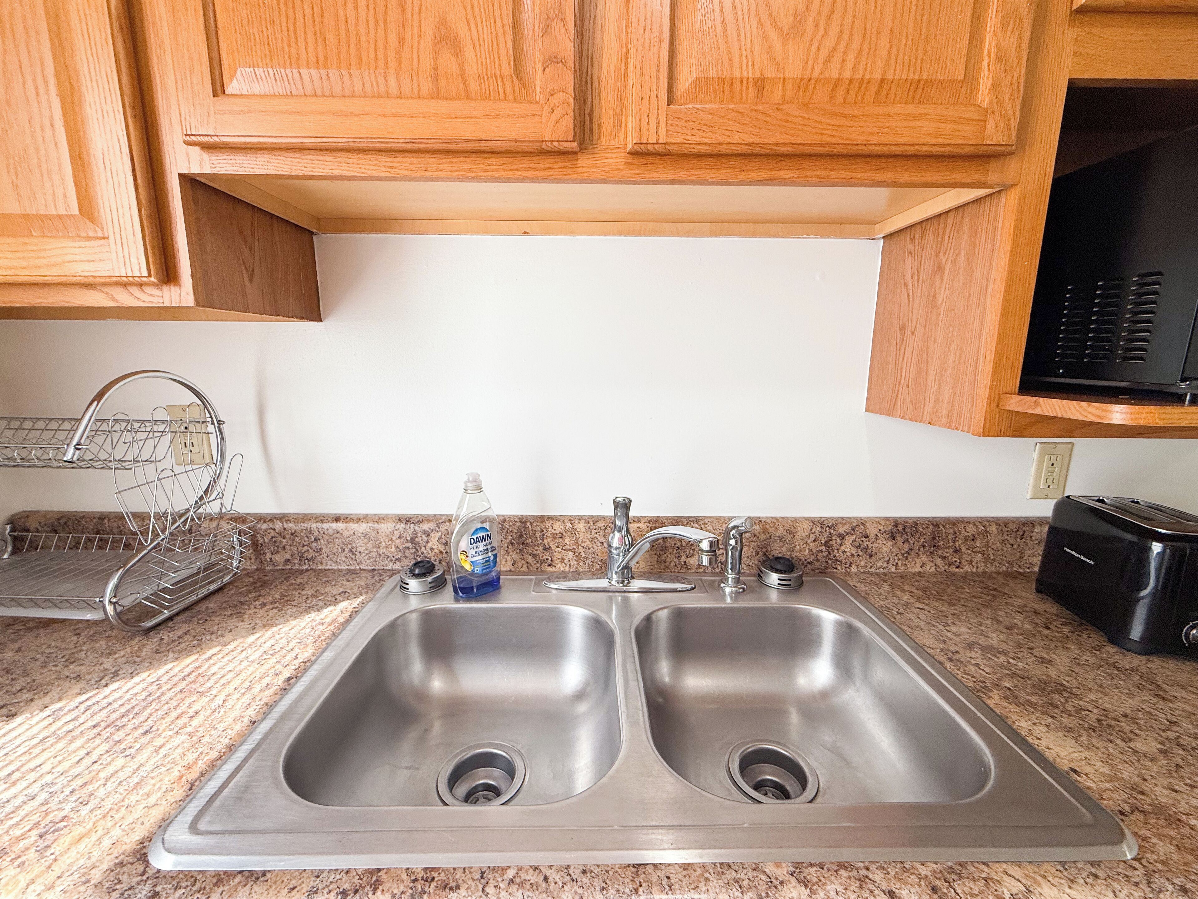 A kitchen with two stainless steel sinks, wooden cabinets, a dish rack, and a black microwave.