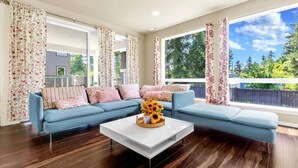 Una sala moderna con un sofá seccional azul, una mesa de centro blanca con un centro de flores y ventanas amplias con vistas a vegetación.