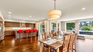 Un comedor amplio con sillas de madera y una mesa preparada para cuatro personas, una cocina con electrodomésticos modernos y taburetes de bar rojos, y ventanas grandes que ofrecen vistas a zonas verdes.