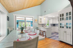 A dining area with a table set for four, a kitchen with white cabinets, and a view of greenery outside.