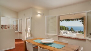 A dining area with a wooden table, white chairs, and a view of the sea through open windows. - New property_Sa Turri: Relax, Infinity Pool, Seaview, enchanting Sunsets (Torre Delle Stelle (Maracalagonis))