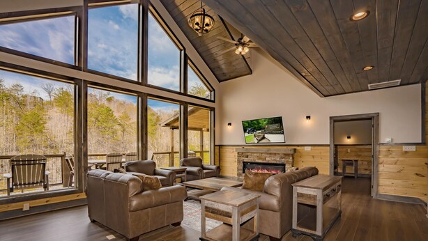 A spacious living room with leather sofas, a fireplace, and large windows overlooking a wooded area.