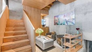 Un intérieur moderne avec un escalier en bois, un banc rembourré de couleur grise, une table d’appoint en bois sur laquelle est posé un vase, et une étagère murale où sont rangés des magazines.