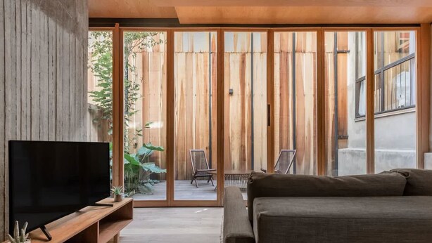 A modern living room with a large flat-screen TV, a sofa, and a sliding glass door leading to an outdoor area with plants and a chair. - Eco Loft with Terrace Garden & Gym Access (Ciudad de México)