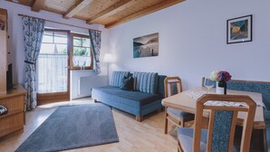 A cozy living room with a blue sofa, wooden dining table, and a view of the outdoors through a window with curtains. - Appartement Plattner - TOP 2 (Zell am See)